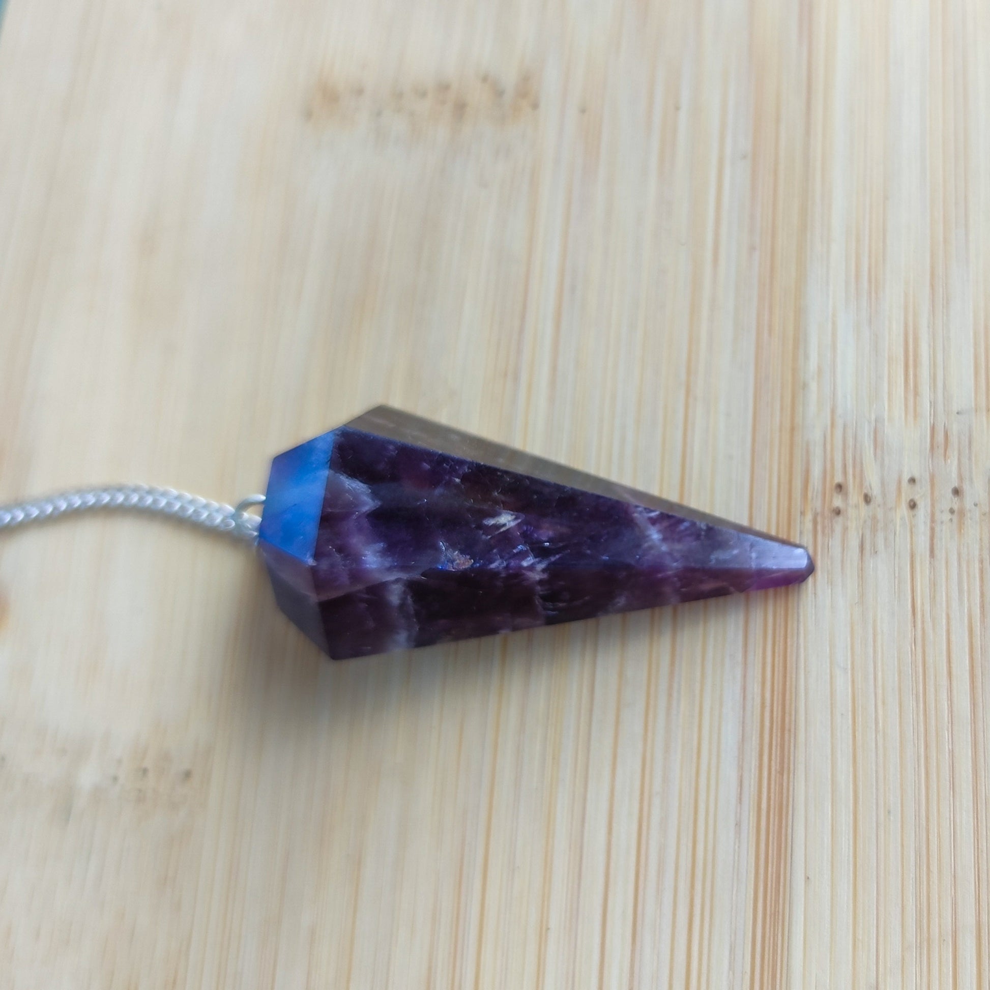 Péndulo de amatista natural violeta de 6 facetas con cadena plateada apoyado de lado.