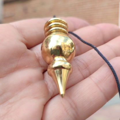 Mano sosteniendo un péndulo de bronce esférico mostrando su tamaño y acabado dorado.