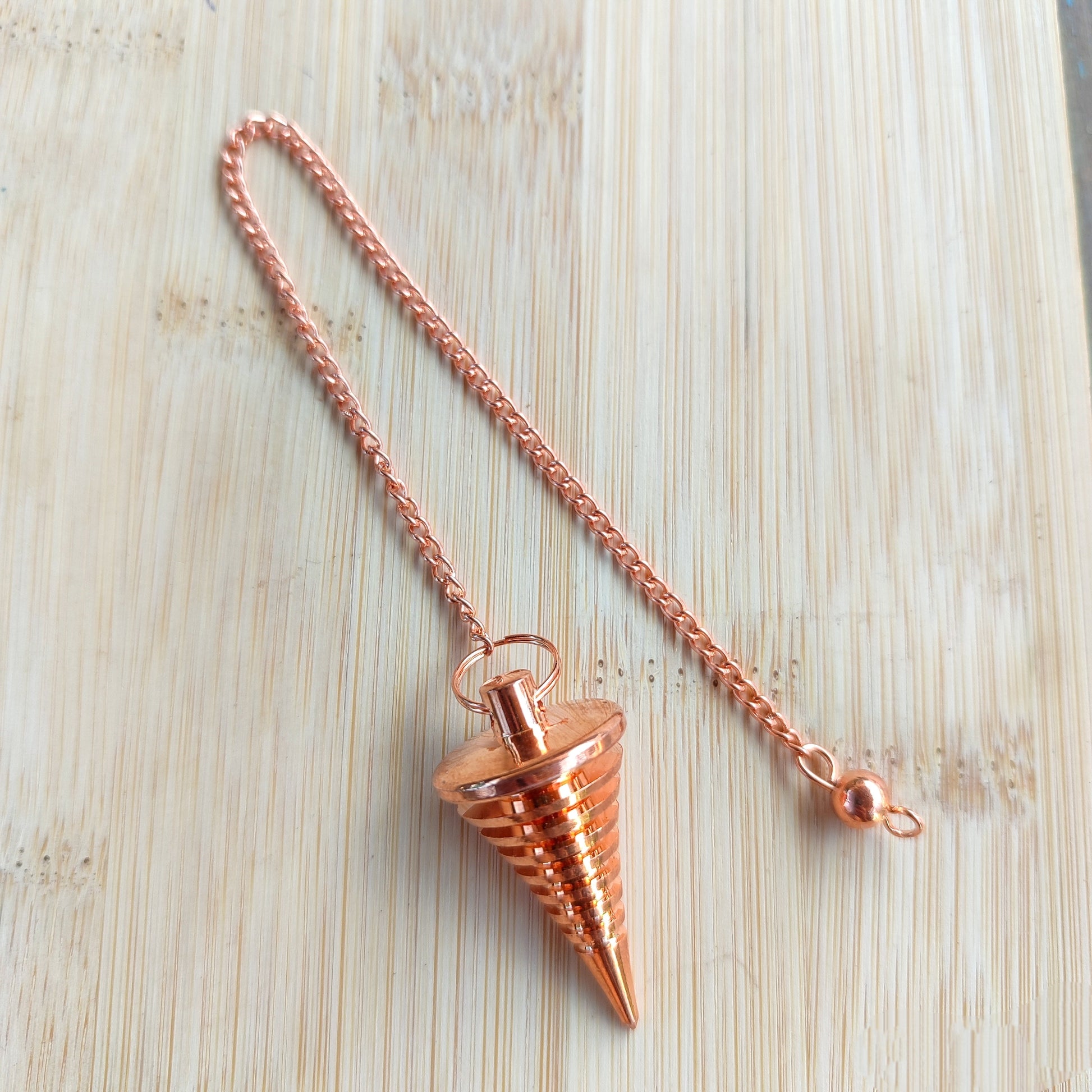 Péndulo de cobre puro con diseño de láminas esféricas superpuestas y cadena sobre madera clara.