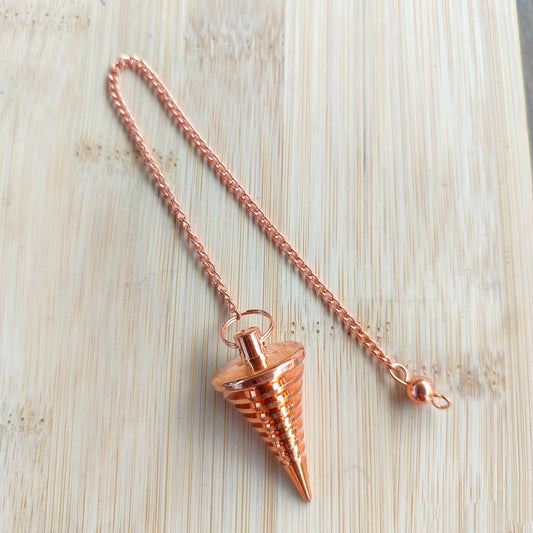 Péndulo de cobre puro con diseño de láminas esféricas superpuestas y cadena sobre madera clara.