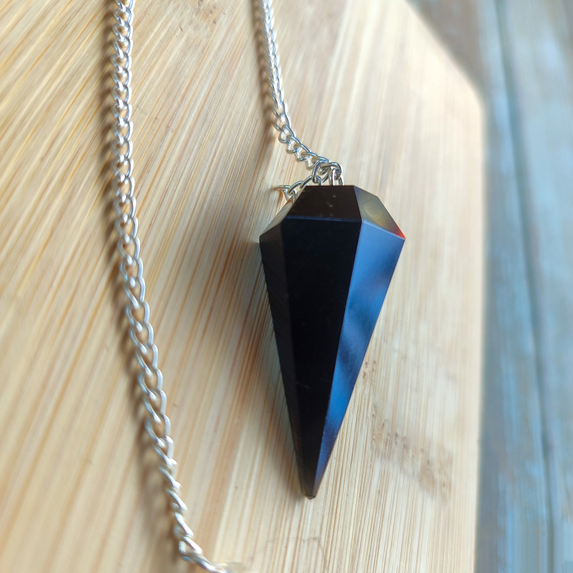 Péndulo de obsidiana negra natural con corte hexagonal de 6 facetas y cadena plateada apoyado sobre madera.